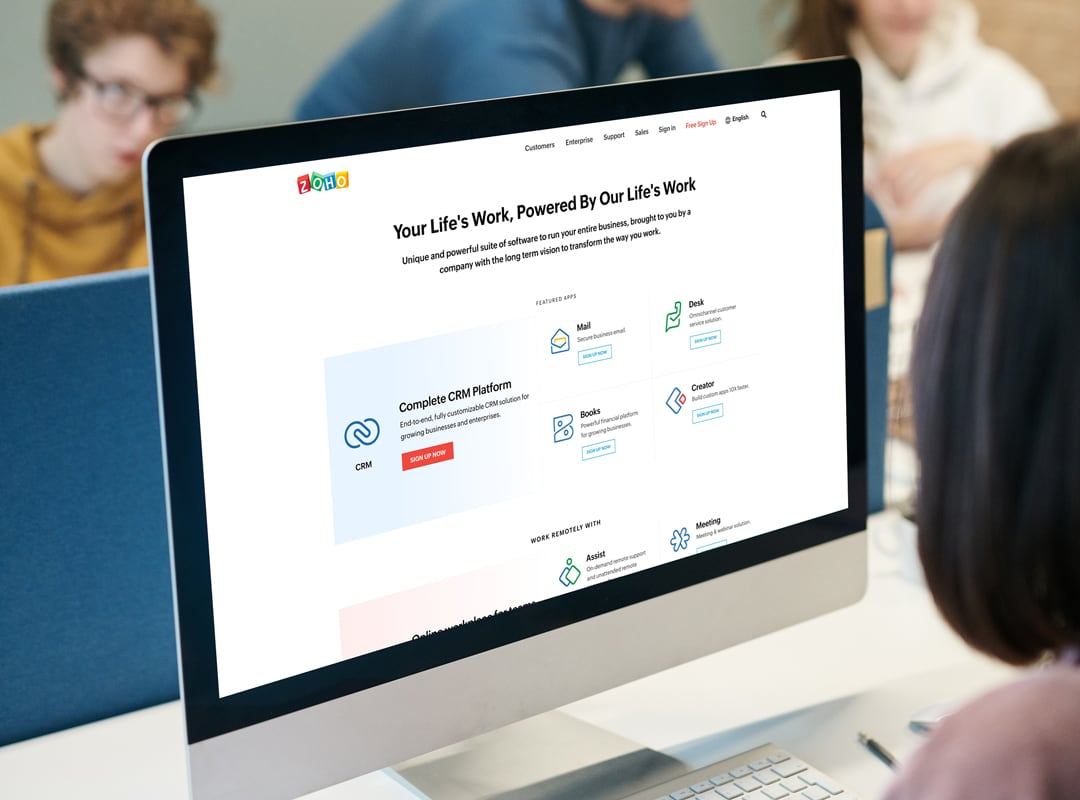 Desktop monitor in a shared office showing the Zoho homepage, with its Complete CRM Platform offer.