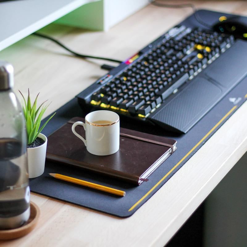 Black Corsair mechanical keyboard with yellow-lit keys on a desk mat beside a notebook, coffee cup and small plant.