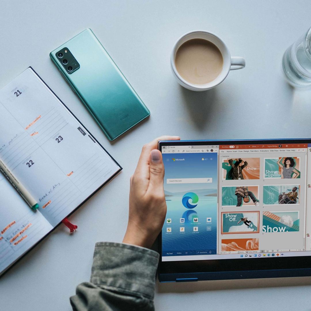 Hand holding a tablet running Microsoft Edge and PowerPoint, beside a phone, coffee and open planner on a desk.