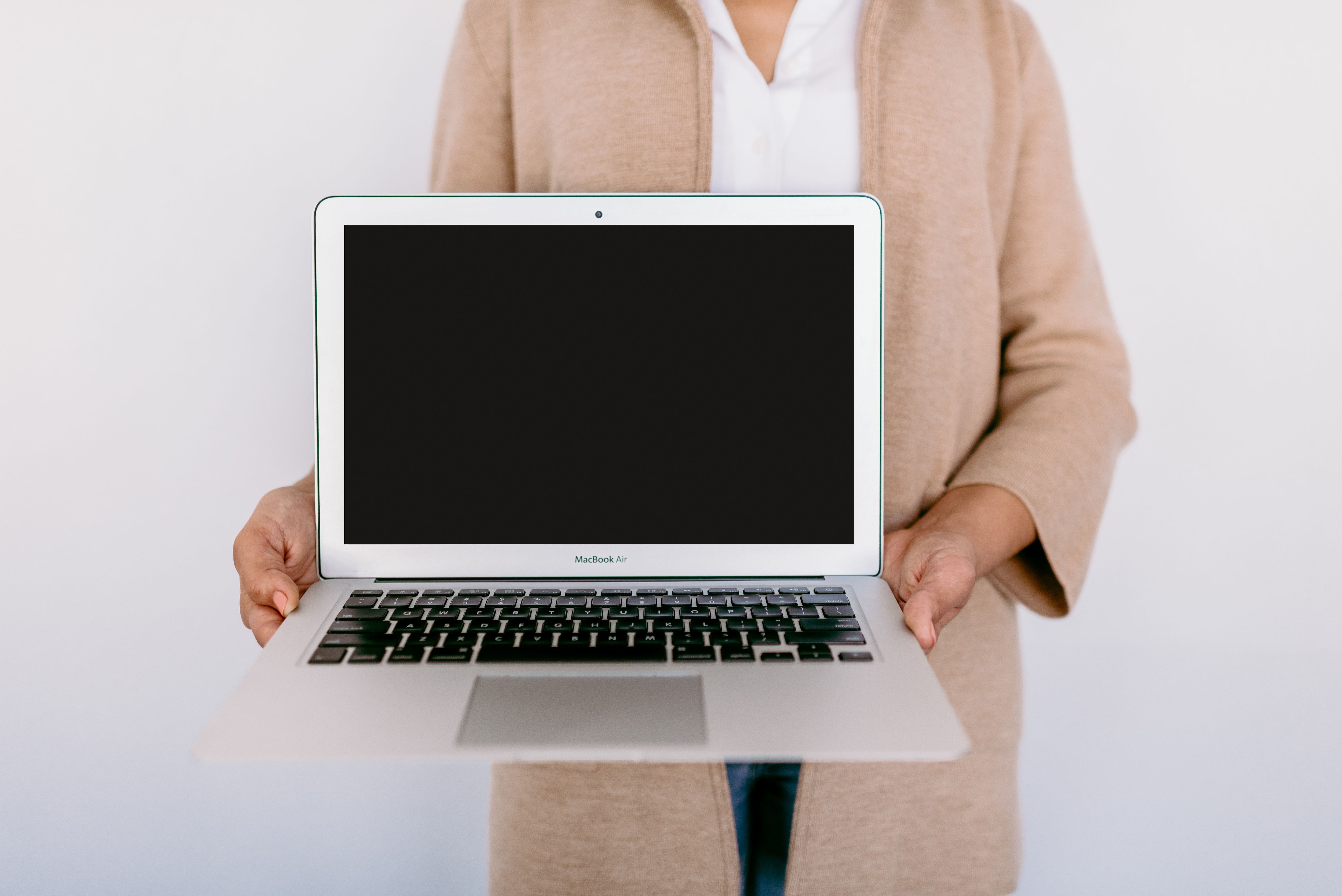 A person dressed in a beige coat and white shirt is holding a MacBook Air with the screen facing forward, displaying a blank screen ready for presentation or to showcase an interface. The background is a plain, light color, providing a neutral setting that emphasizes the laptop.