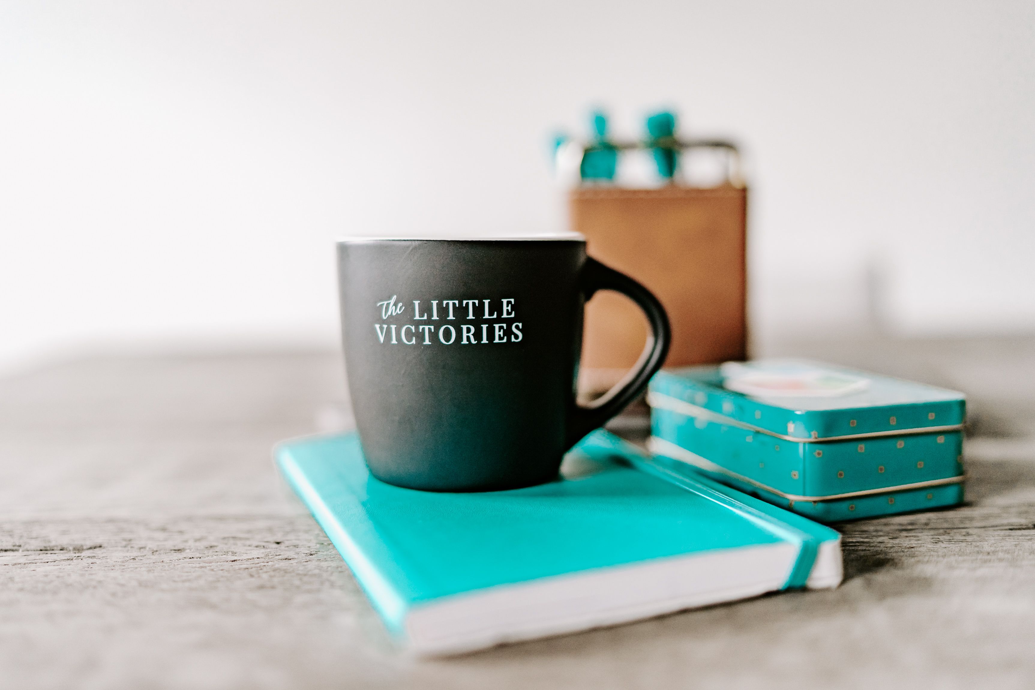 A black matte mug with the inscription "The LITTLE VICTORIES" sits on top of a bright teal notebook, with a small, matching teal tin box adorned with golden dots in the background. The setting suggests a workspace with a focus on celebrating small achievements, possibly in a home or casual office environment.