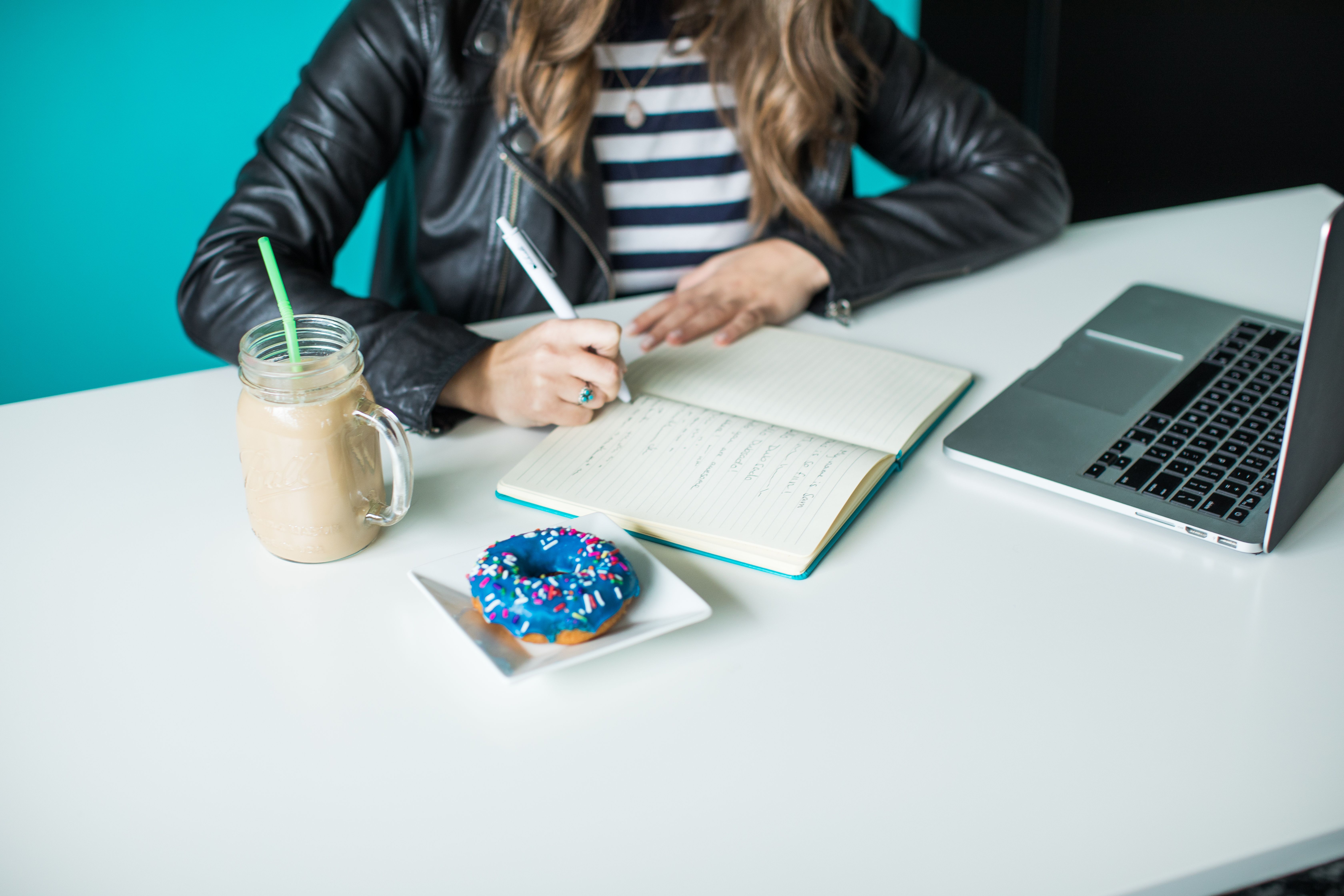 A person is seated at a white desk, writing in a notebook, with a laptop to the right and a frosted doughnut topped with colorful sprinkles to the left. In front of them is a mason jar with a creamy beverage and a green straw, suggesting a casual work or study setting. The teal backdrop adds a vibrant contrast to the scene, which evokes productivity with a touch of indulgence.