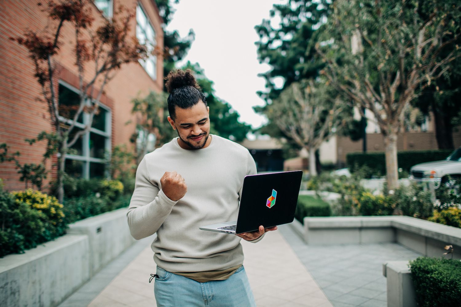 A man in a light sweater and jeans stands outside, triumphantly raising his fist while looking at his laptop with a colorful logo on the back. He appears to be celebrating a success or victory that he has just discovered or achieved online.