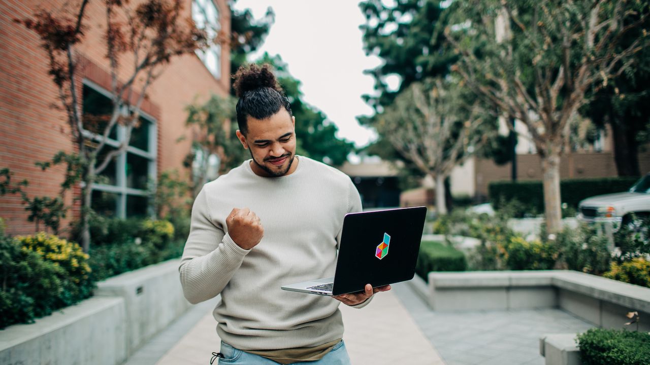 A man in a light sweater and jeans stands outside, triumphantly raising his fist while looking at his laptop with a colorful logo on the back. He appears to be celebrating a success or victory that he has just discovered or achieved online.