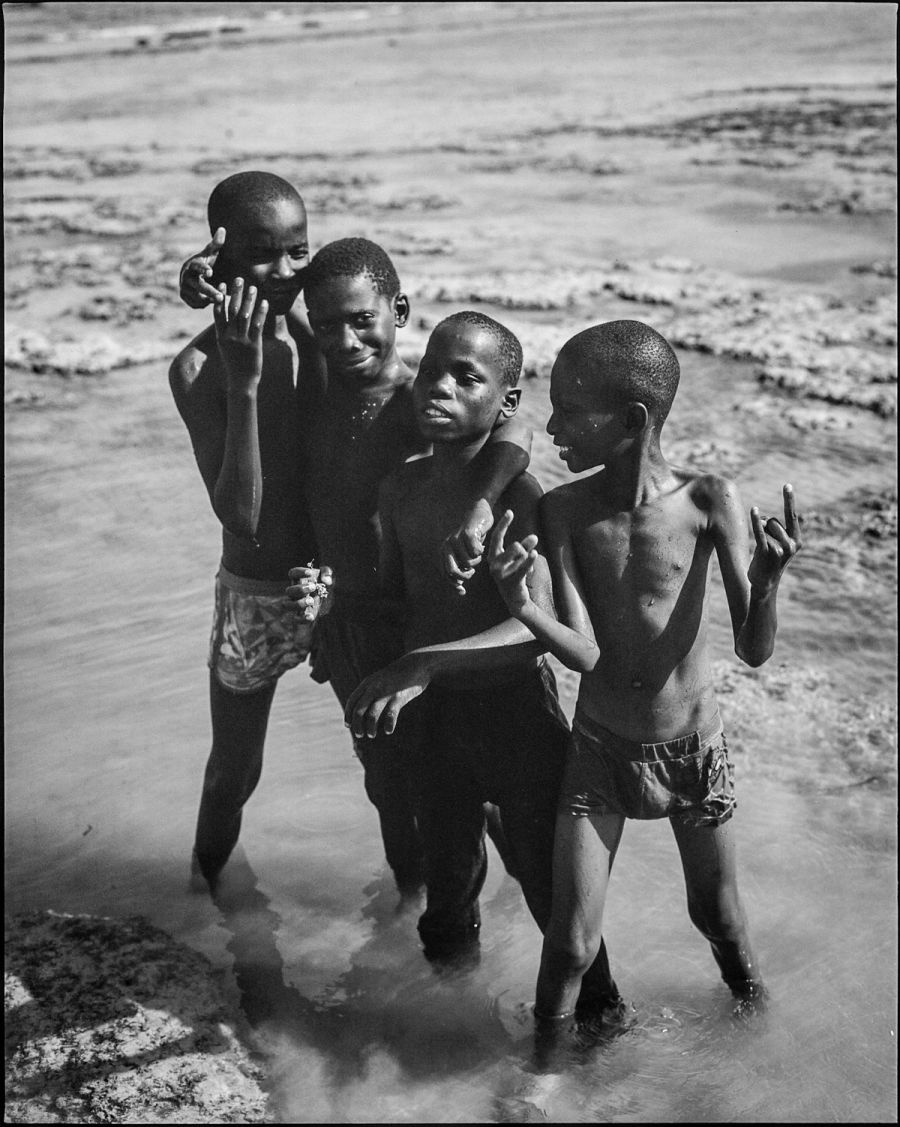 Four boys stand together in shallow water, smiling and making hand gestures, film photograph by Nicolò Pudel.