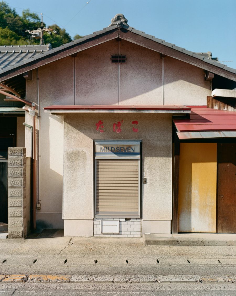 A shuttered storefront with Japanese characters and "MIDSEVEN" signage on a weathered concrete building under clear sky, film photograph by Nicolò Pudel