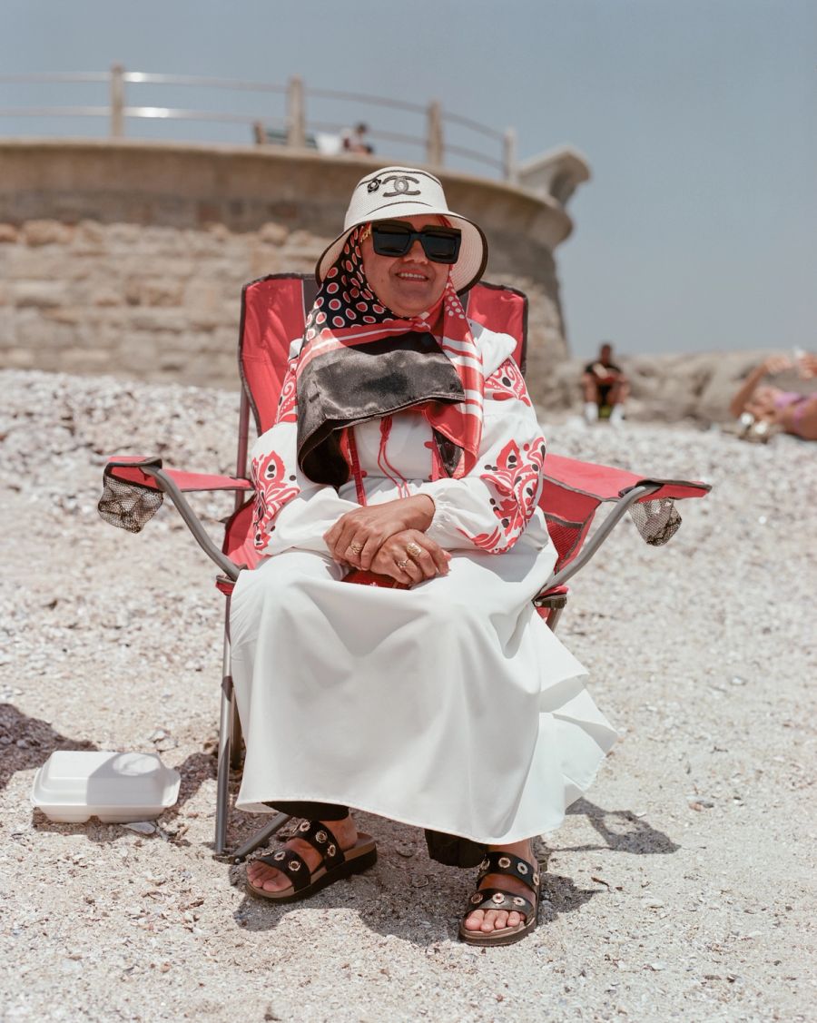 Woman in a bucket hat and sunglasses seated on a folding chair on a pebble beach. Film photograph by Nicolò Pudel