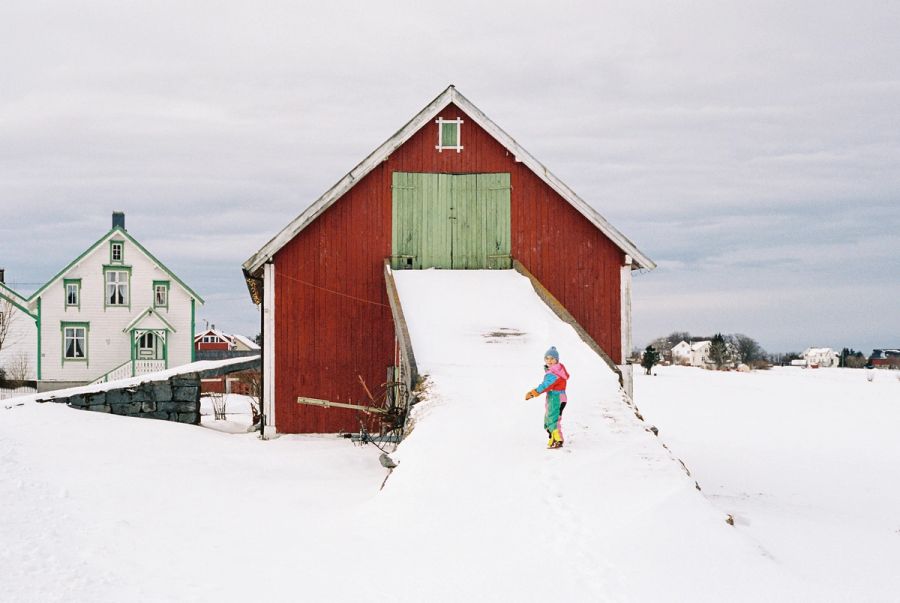 Child in a colourful snowsuit on the snowed-in ramp of a red barn, Norway. Film photograph by Nicolò Pudel