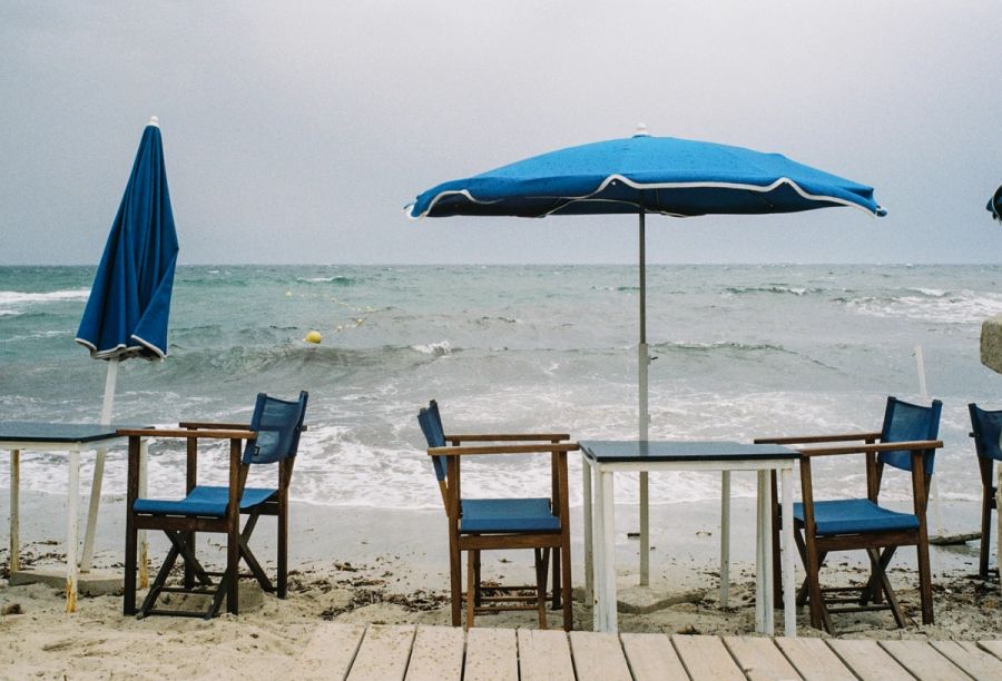 Blue umbrellas and chairs on a wooden beach deck under grey sky. Film photograph by Nicolò Pudel