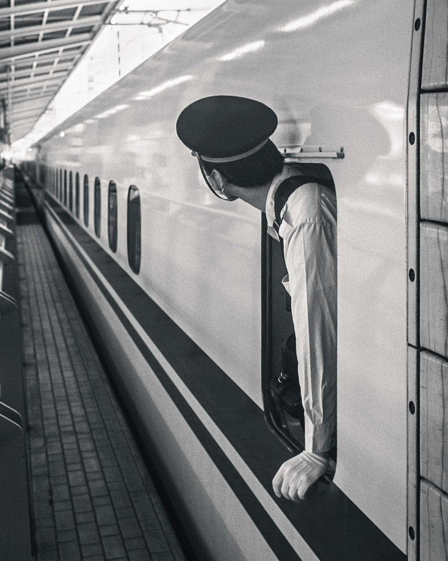 Train conductor in uniform standing in doorway of passenger train at station platform, film photograph by Nicolò Pudel.
