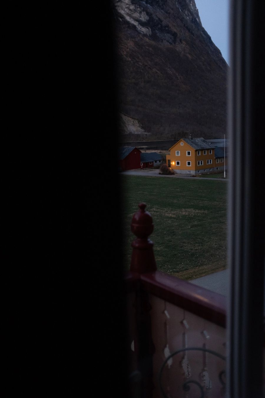 Yellow farmhouse at dusk beneath a dark mountainside, seen from a window, Norway. Film photograph by Nicolò Pudel