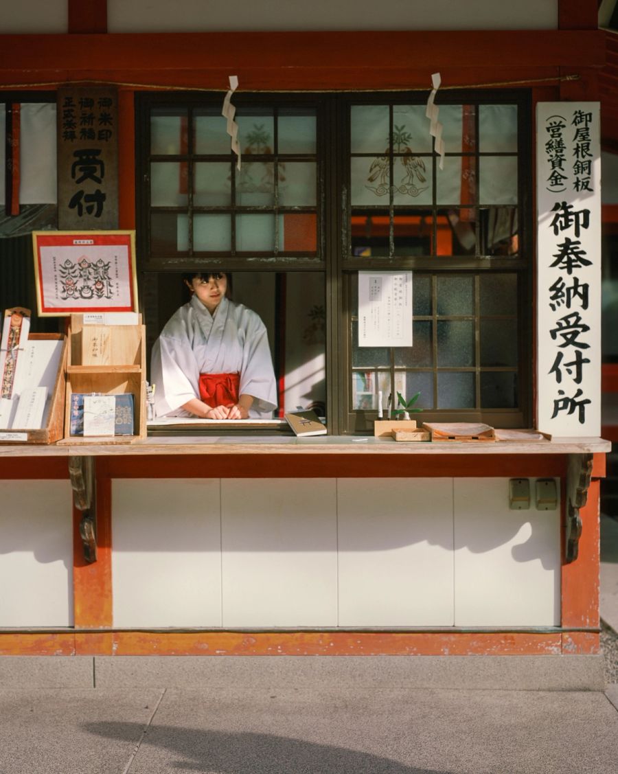 A woman in traditional Japanese clothing sits at a temple donation window with Japanese signage and religious offerings displayed, film photograph by Nicolò Pudel.