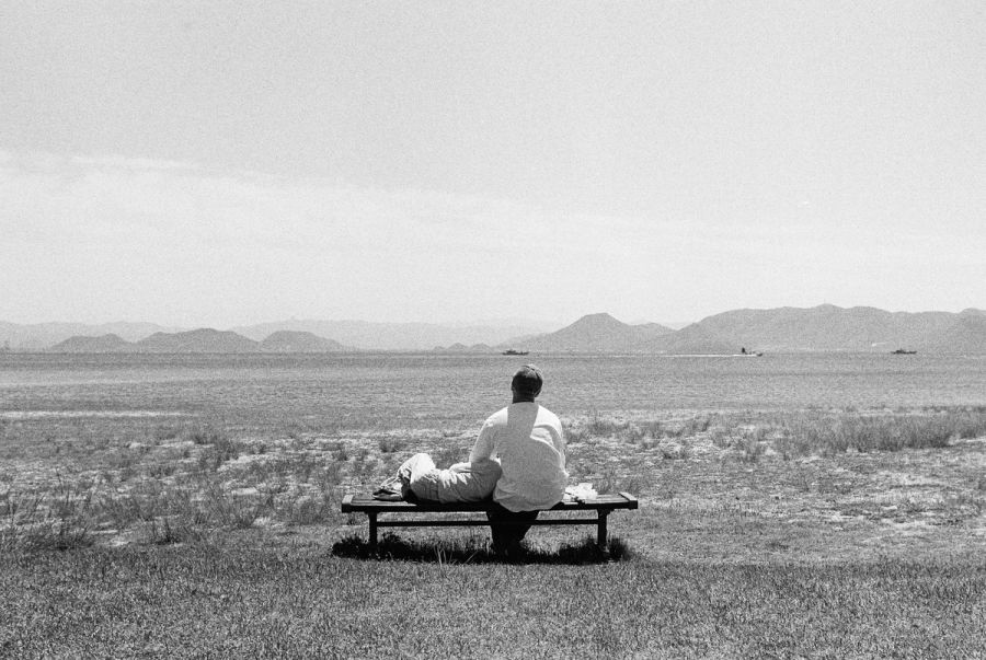 A person sits alone on a bench facing a vast bay with distant mountains, film photograph by Nicolò Pudel.