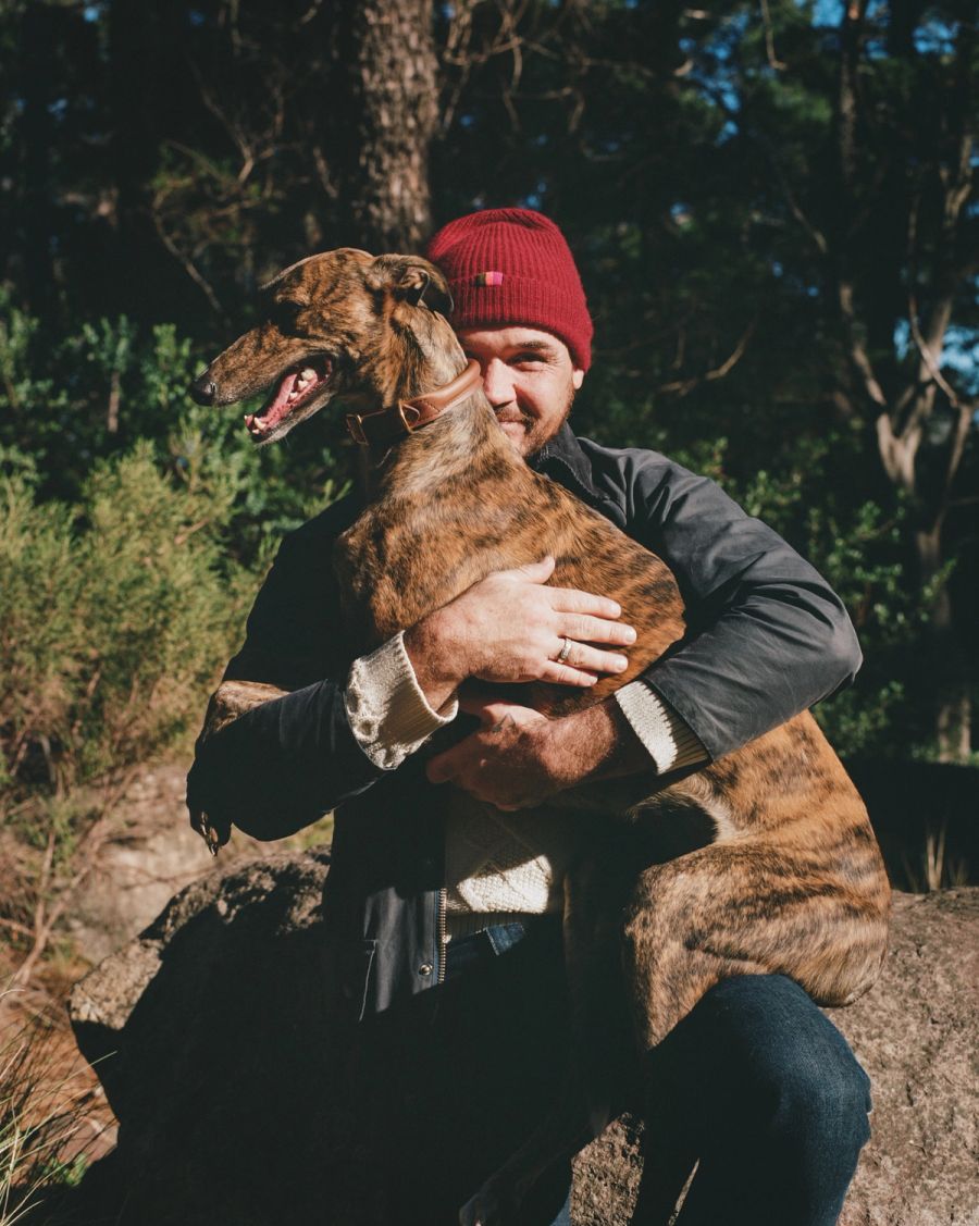 Man in a red beanie hugging a brindle greyhound in the forest, Deer Park, Cape Town. Film photograph by Nicolò Pudel
