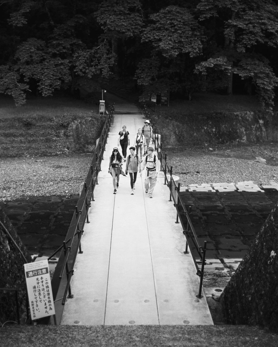 A group of hikers walks across a wooden bridge through a forested ravine, film photograph by Nicolò Pudel.
