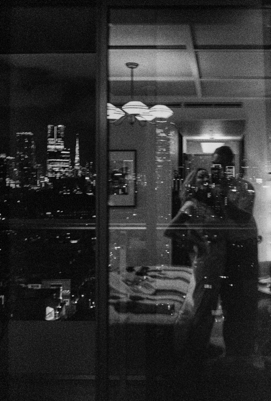 A woman in an apartment interior framed by windows overlooking a nighttime city skyline, film photograph by Nicolò Pudel.