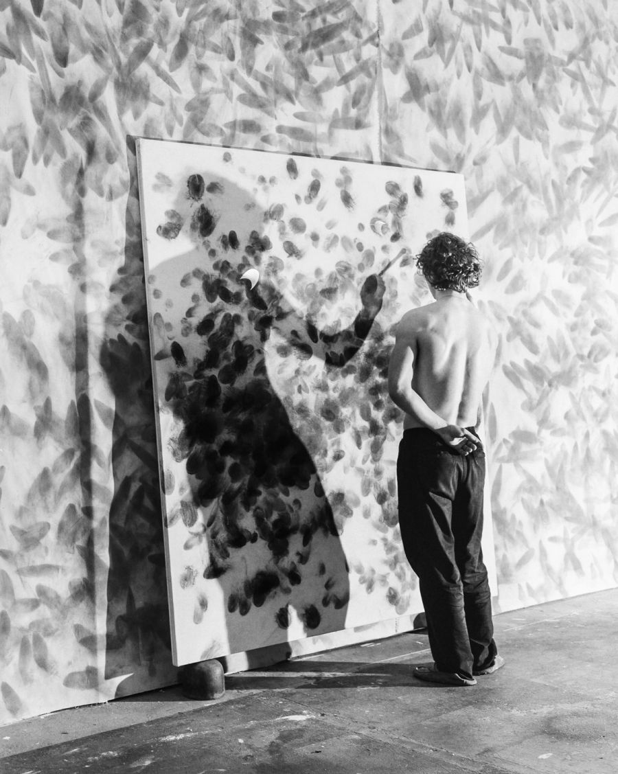 A shirtless man stands before a large panel covered with dark handprints against a leafy backdrop, film photograph by Nicolò Pudel