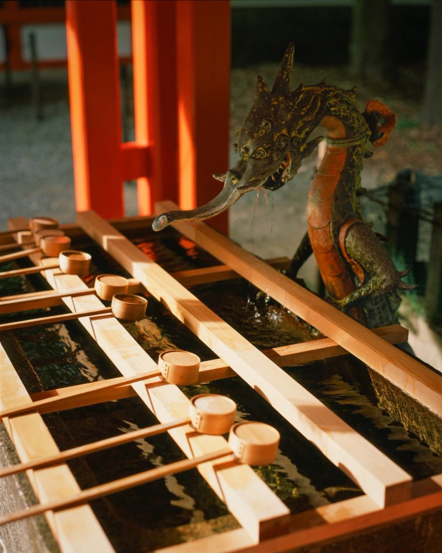 Ornate brass dragon spout with wooden ladles positioned over channels in a Japanese temple purification fountain, film photograph by Nicolò Pudel