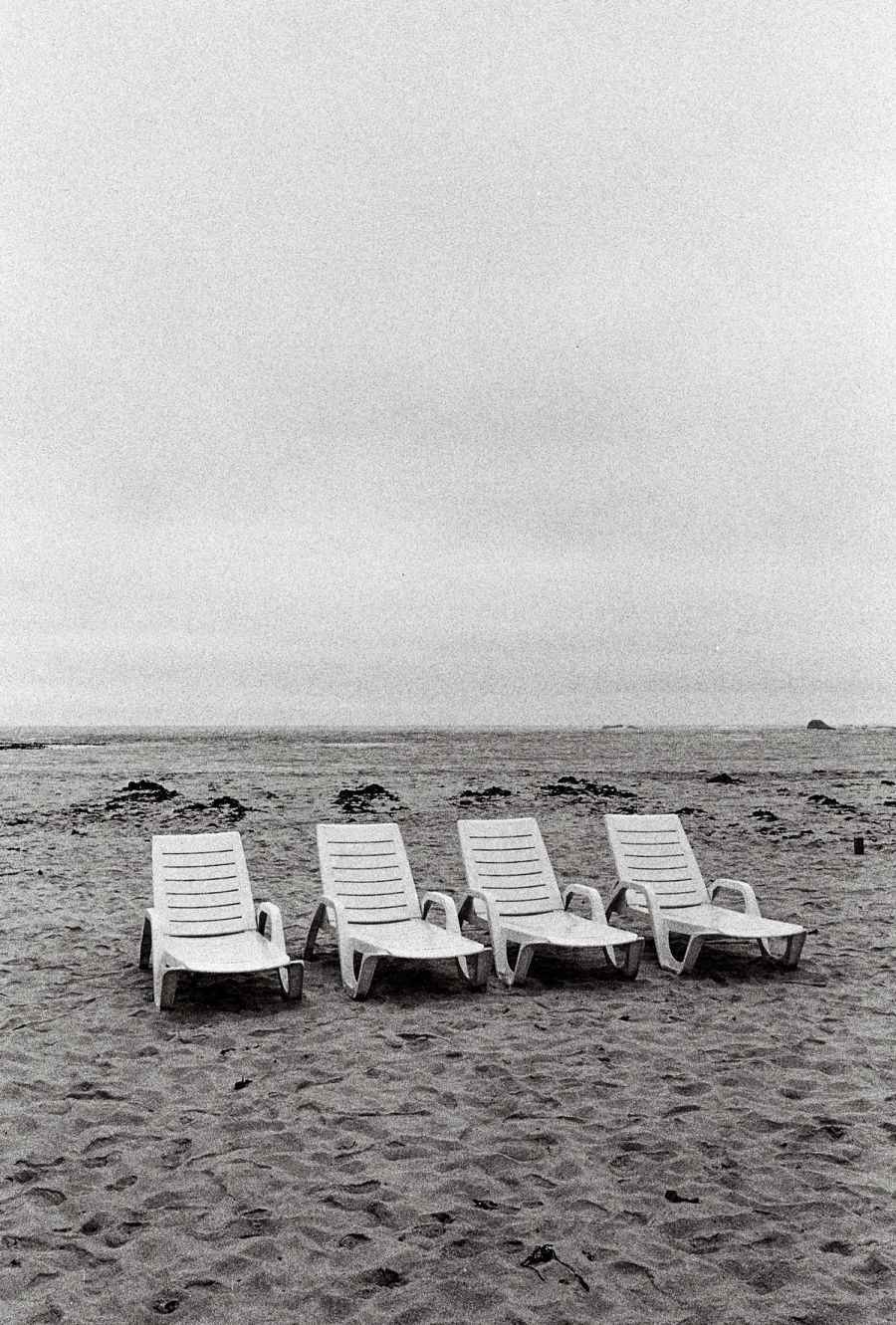 Four empty white sun loungers on a grey overcast beach, black and white. Film photograph by Nicolò Pudel