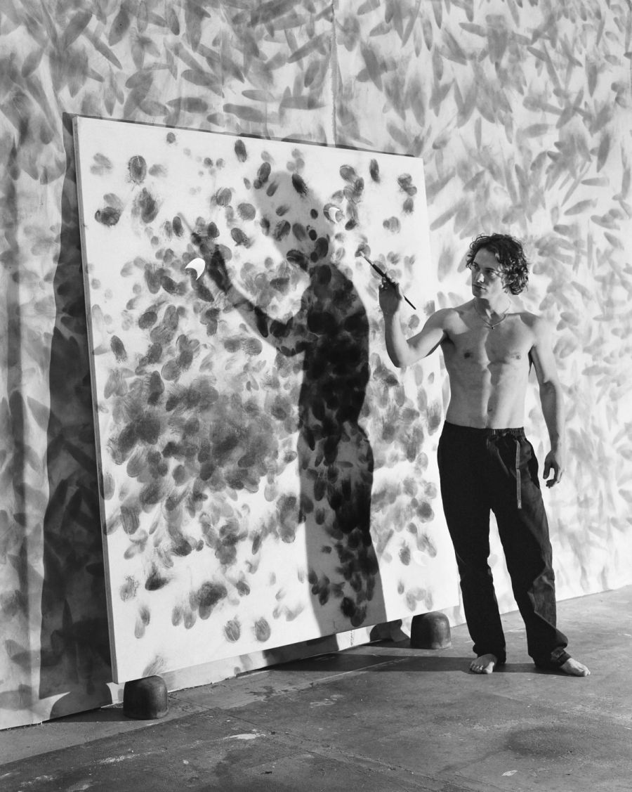 A shirtless man holds a brush while standing beside a large canvas covered with handprints, film photograph by Nicolò Pudel.