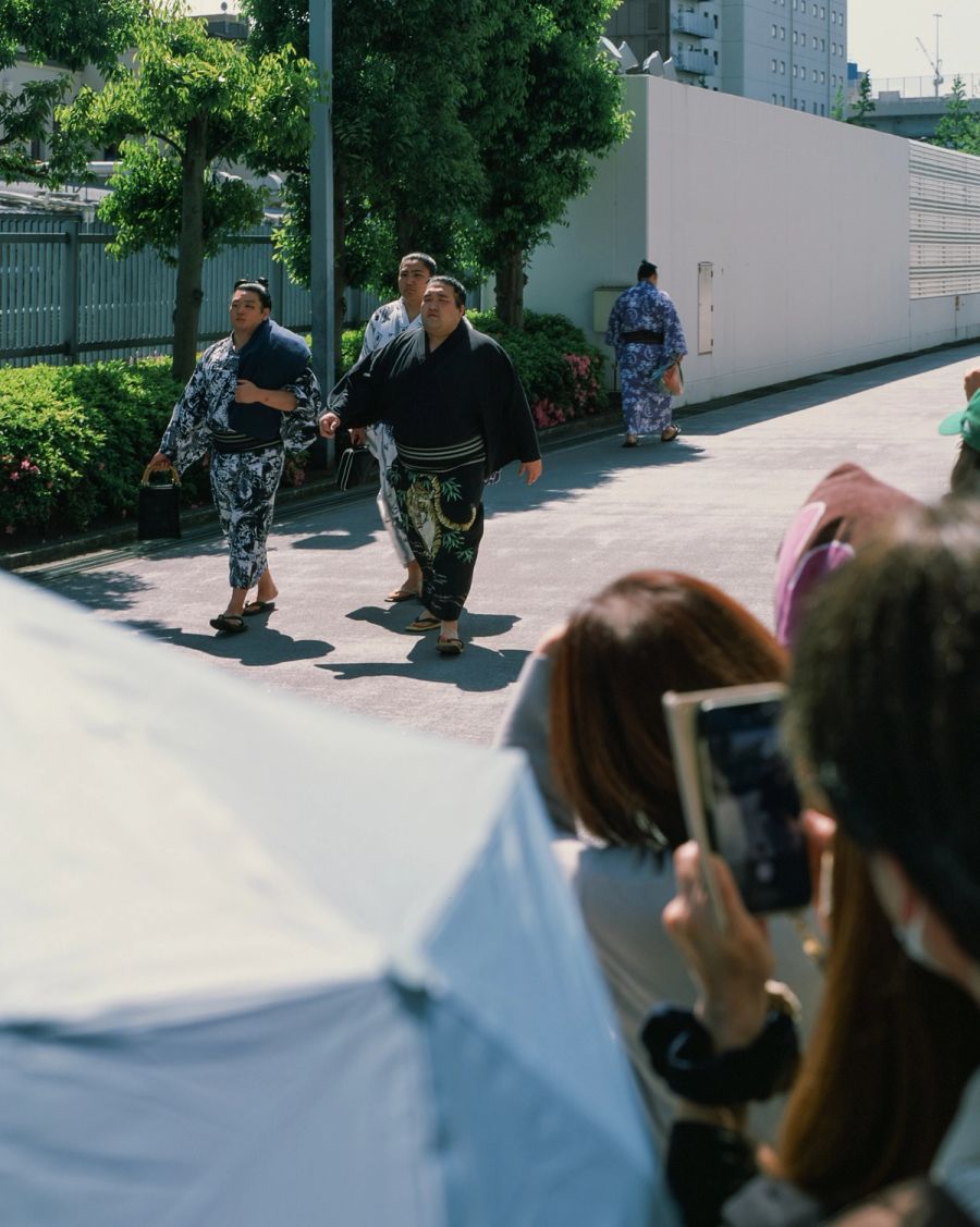 People in traditional kimono walk across a sunny urban plaza while a woman in the foreground photographs them, film photograph by Nicolò Pudel.