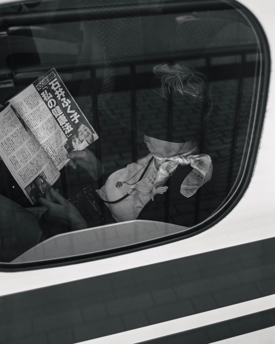 A person reads a Japanese newspaper inside a car, viewed through the side window, film photograph by Nicolò Pudel.