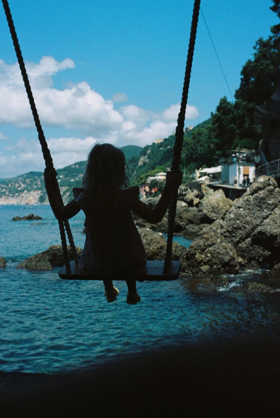 Child on a rope swing looking out over a rocky cove, Ligurian coast, Italy. Film photograph by Nicolò Pudel