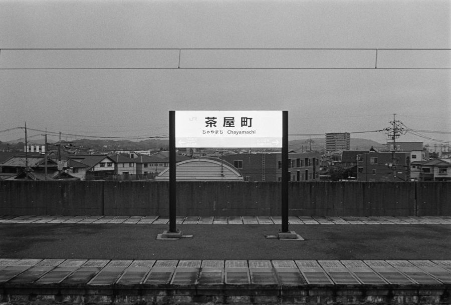 A train station sign reading "Chayamachi" stands on an empty platform overlooking a Japanese town, film photograph by Nicolò Pudel.