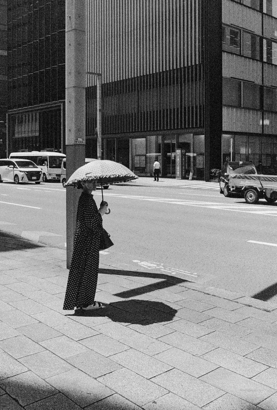 A woman with a polka dot umbrella stands on an urban street beside modernist concrete buildings and parked cars, film photograph by Nicolò Pudel