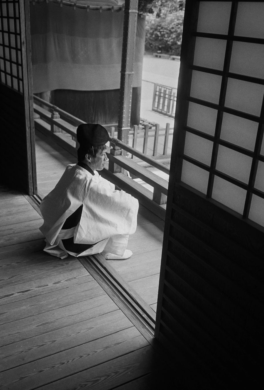 A man in traditional Japanese clothing sits on a wooden veranda looking out toward a garden, film photograph by Nicolò Pudel.