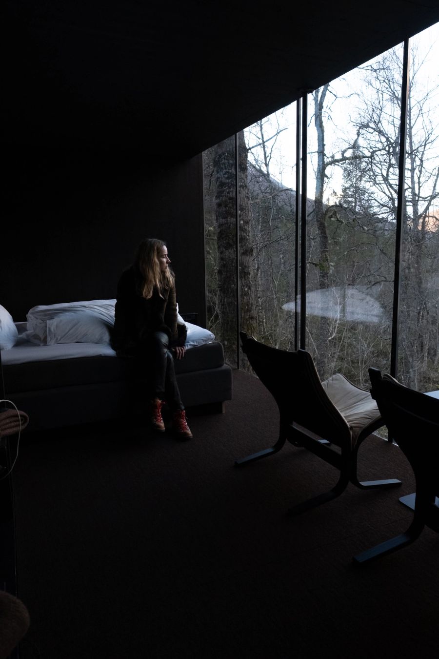 Woman seated on a bed in a dark cabin facing a wall of bare-forest windows, Norway. Film photograph by Nicolò Pudel
