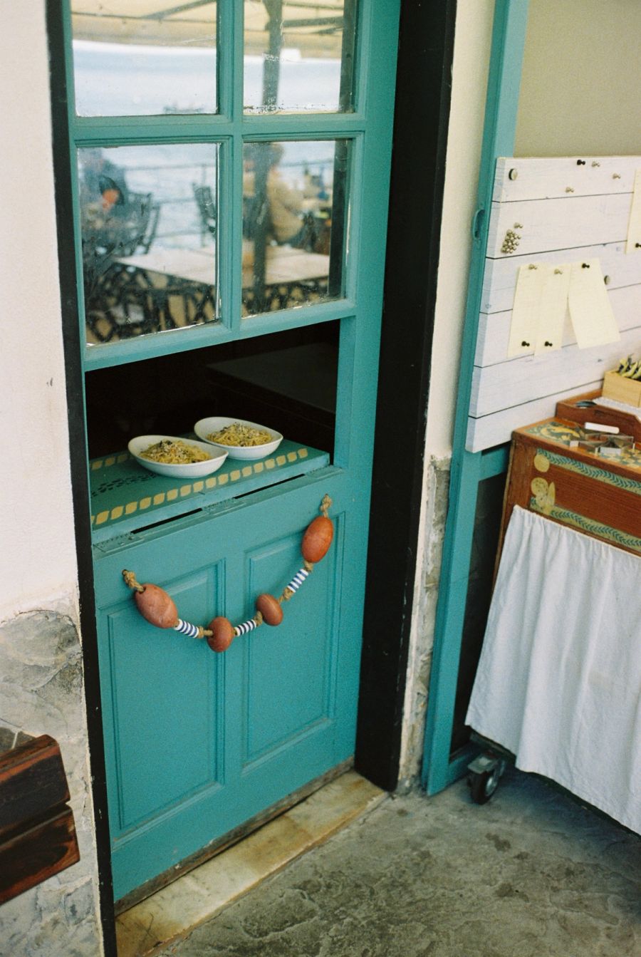 Turquoise wooden door of a seaside restaurant with bowls of pasta on the counter, Italy. Film photograph by Nicolò Pudel