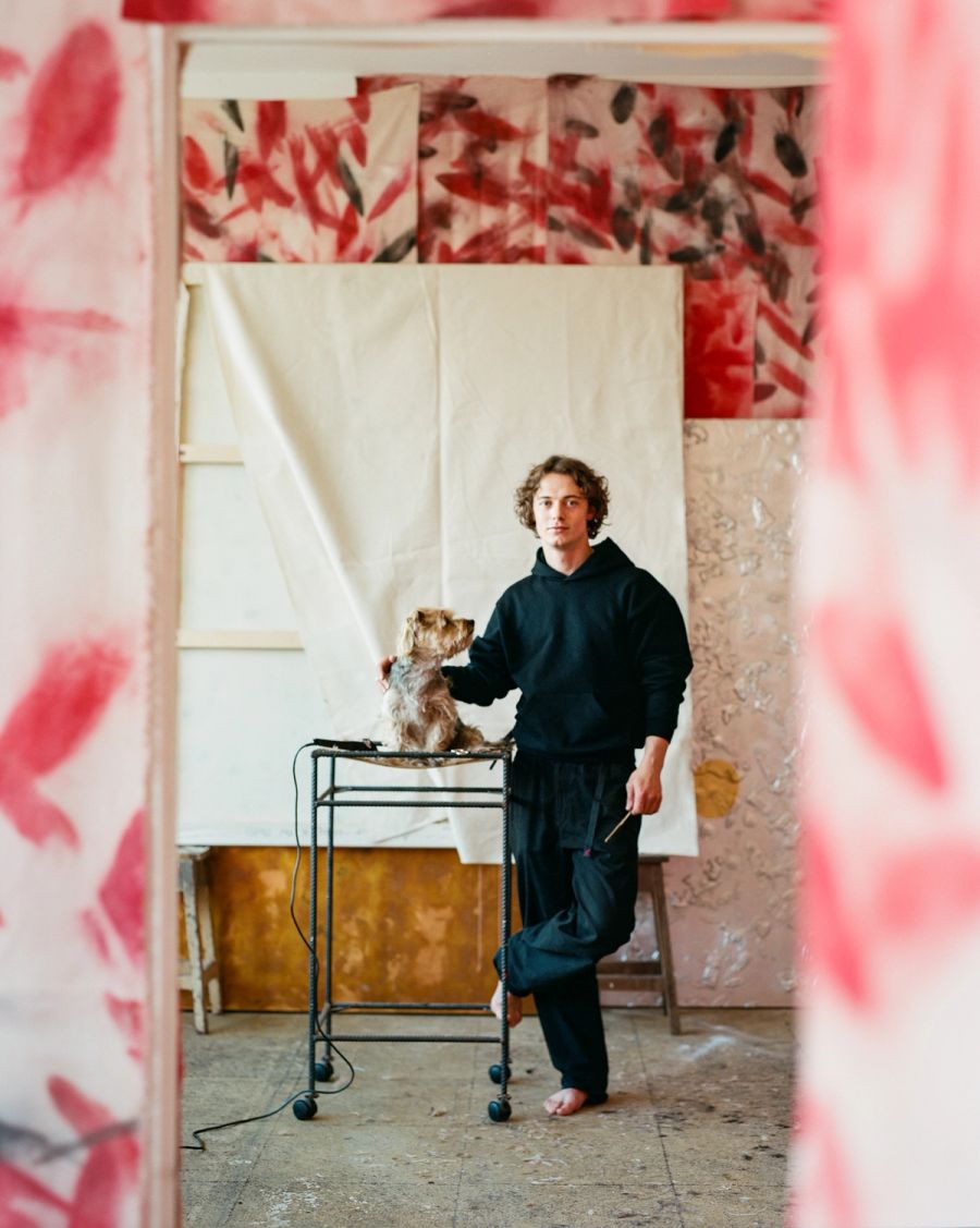 A man in a black hoodie stands beside a small dog on a metal cart in a sparse interior, framed through a pink floral doorway, film photograph by Nicolò Pudel.