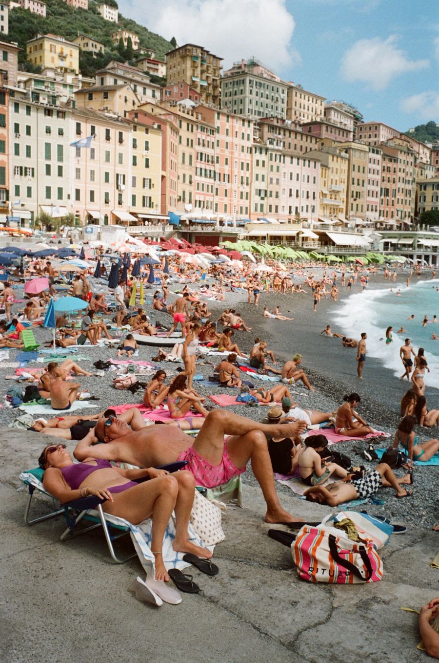 Crowded pebble beach below tall pastel houses on the Italian coast, photographed on film. Film photograph by Nicolò Pudel