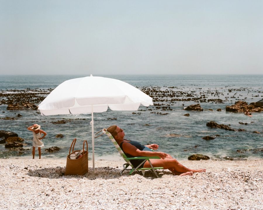White parasol over a sunbather on a rocky beach with kelp in the water, Cape Town. Film photograph by Nicolò Pudel