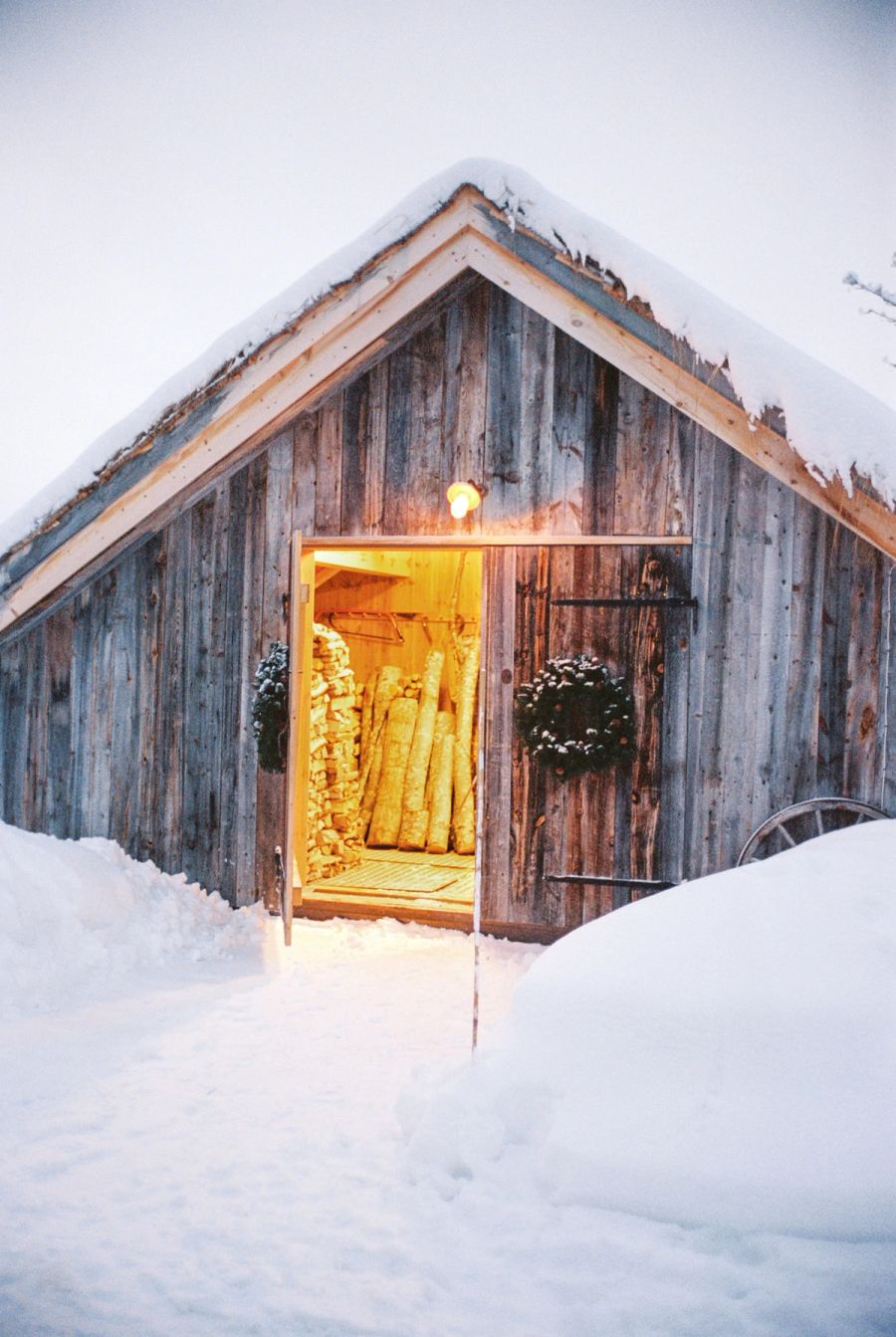 Snow-covered timber barn at dusk with firewood stacked in the lit doorway, Norway. Film photograph by Nicolò Pudel