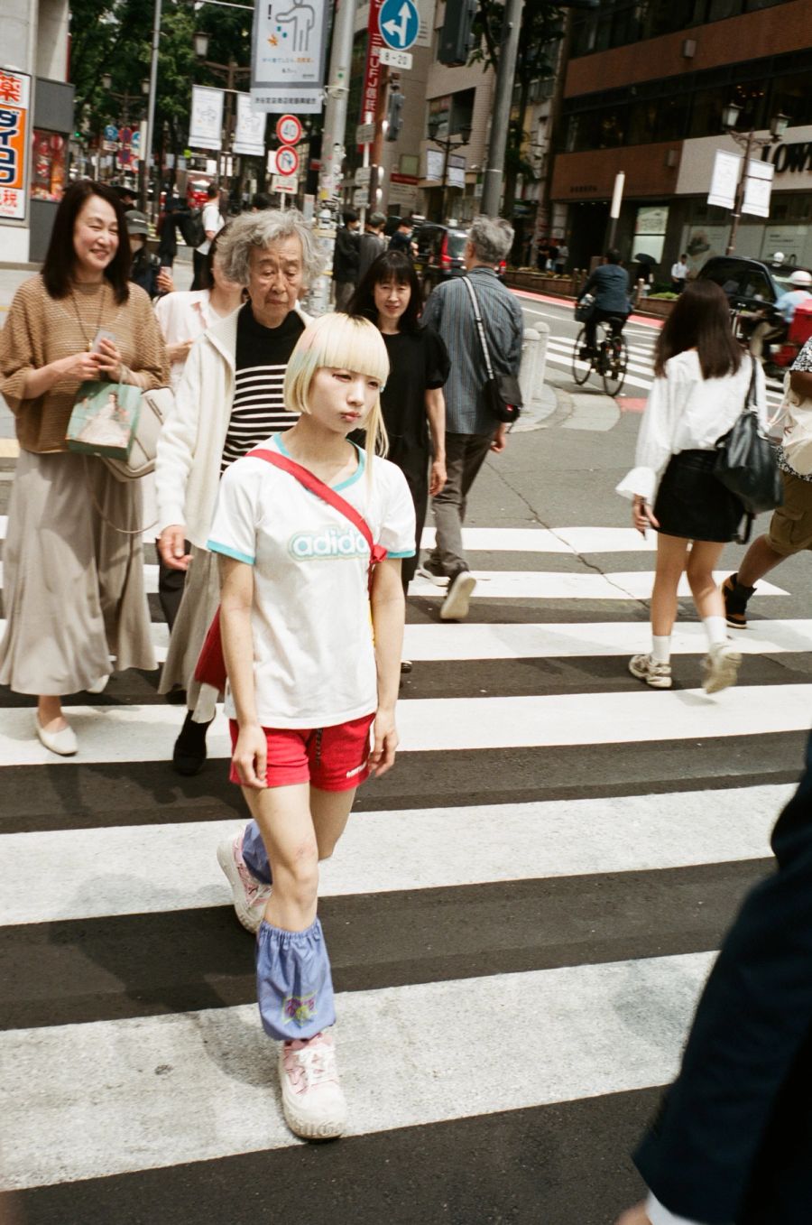 A young woman with blonde bangs crosses a pedestrian crosswalk in a busy urban street lined with Japanese signage, film photograph by Nicolò Pudel.