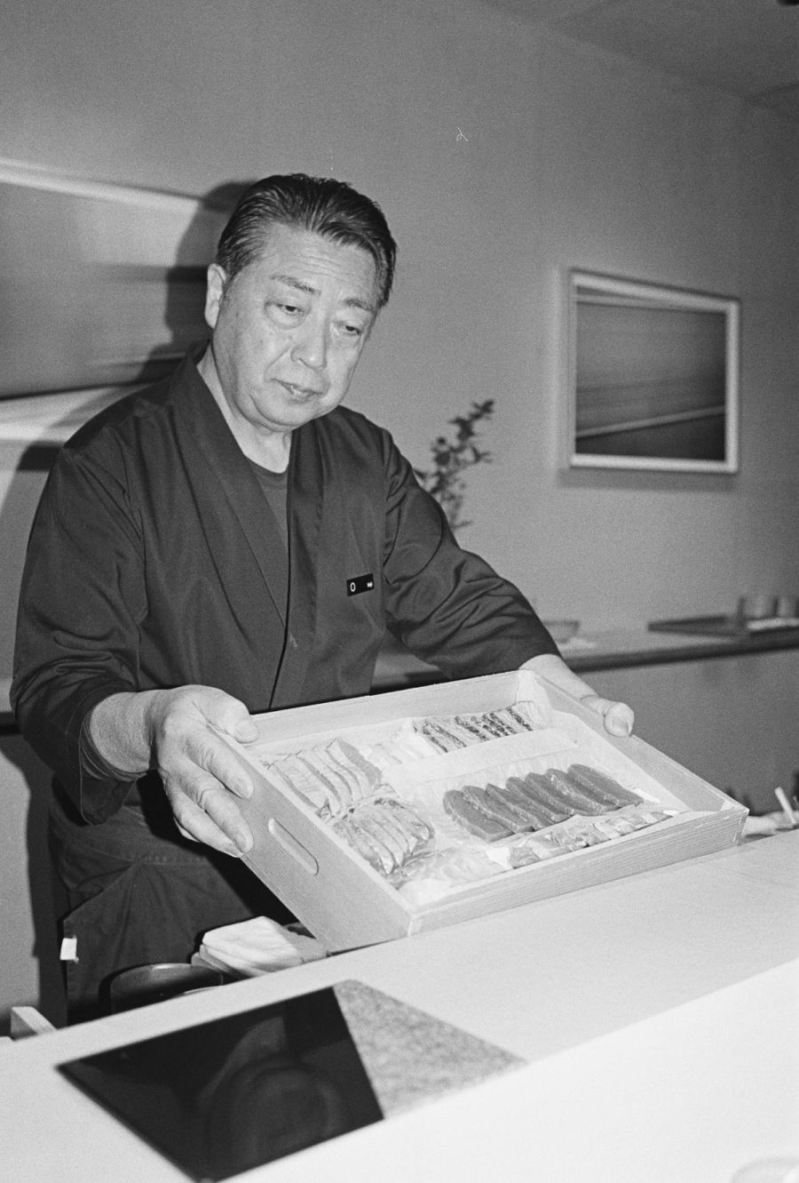 A man examines an open box of preserved food samples at a table indoors, film photograph by Nicolò Pudel.