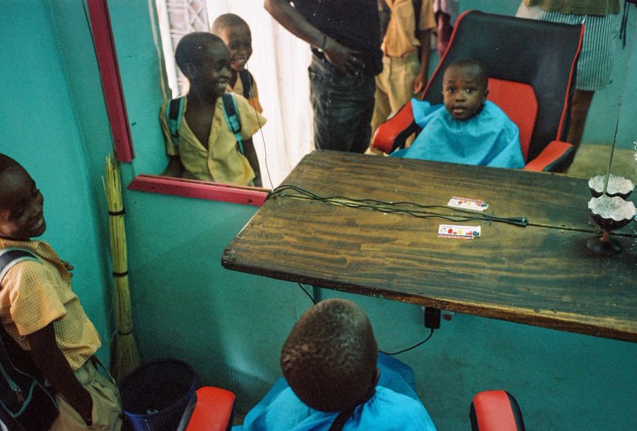 Boy in a barber's cape watched by other children in a teal barbershop, Msambweni, Kenya. Film photograph by Nicolò Pudel