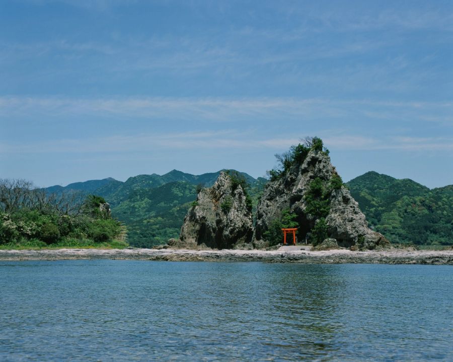 Rocky outcrop with red torii gate on beach, forested mountains beyond, film photograph by Nicolò Pudel