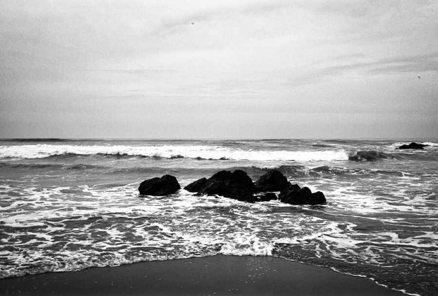 Waves breaking over dark rocks on a grey shore, black and white. Film photograph by Nicolò Pudel