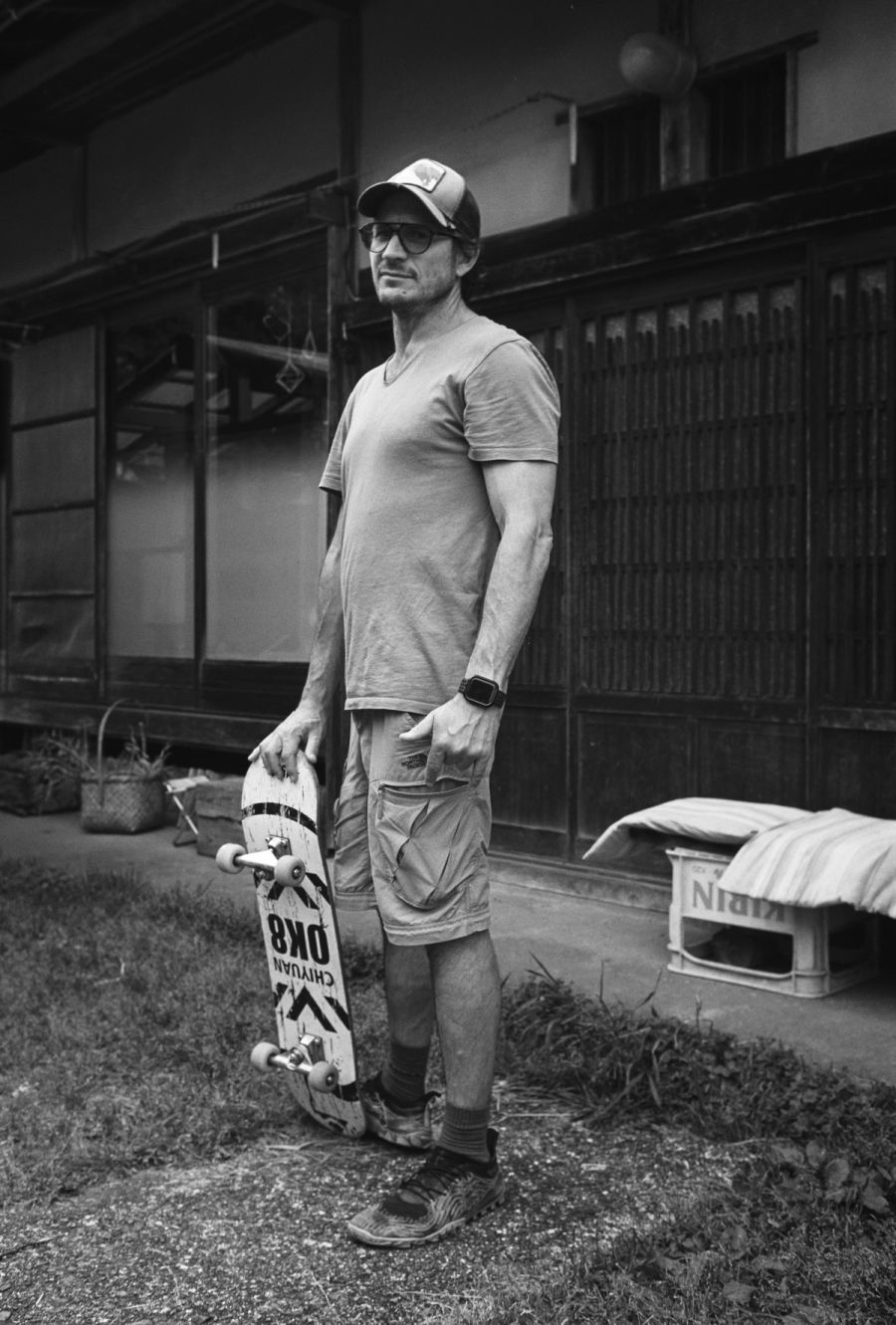 A man wearing a cap and sunglasses holds a skateboard outside a residential building, film photograph by Nicolò Pudel