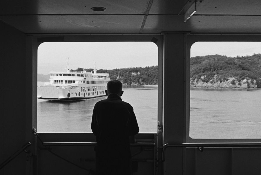 A man stands inside a ferry looking out at a passing riverboat and forested riverbank through large windows, film photograph by Nicolò Pudel.