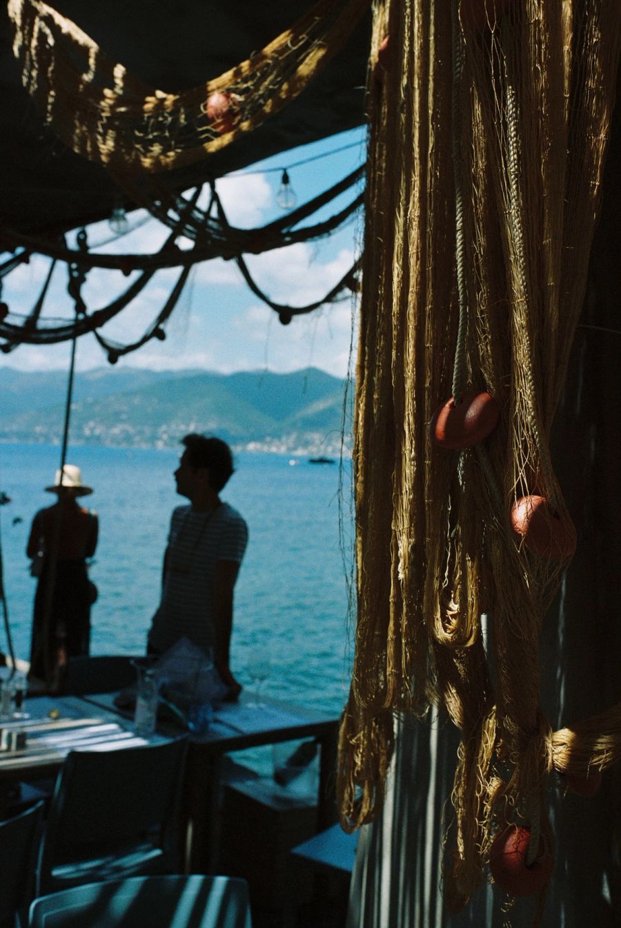 Man at a seaside restaurant table behind hanging fishing nets, Ligurian coast, Italy. Film photograph by Nicolò Pudel