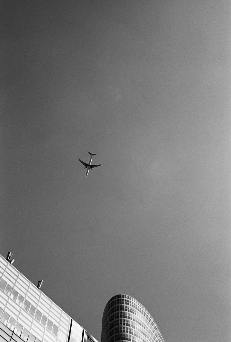 Airplane flying overhead above modern cylindrical and rectangular buildings in an urban setting, film photograph by Nicolò Pudel.