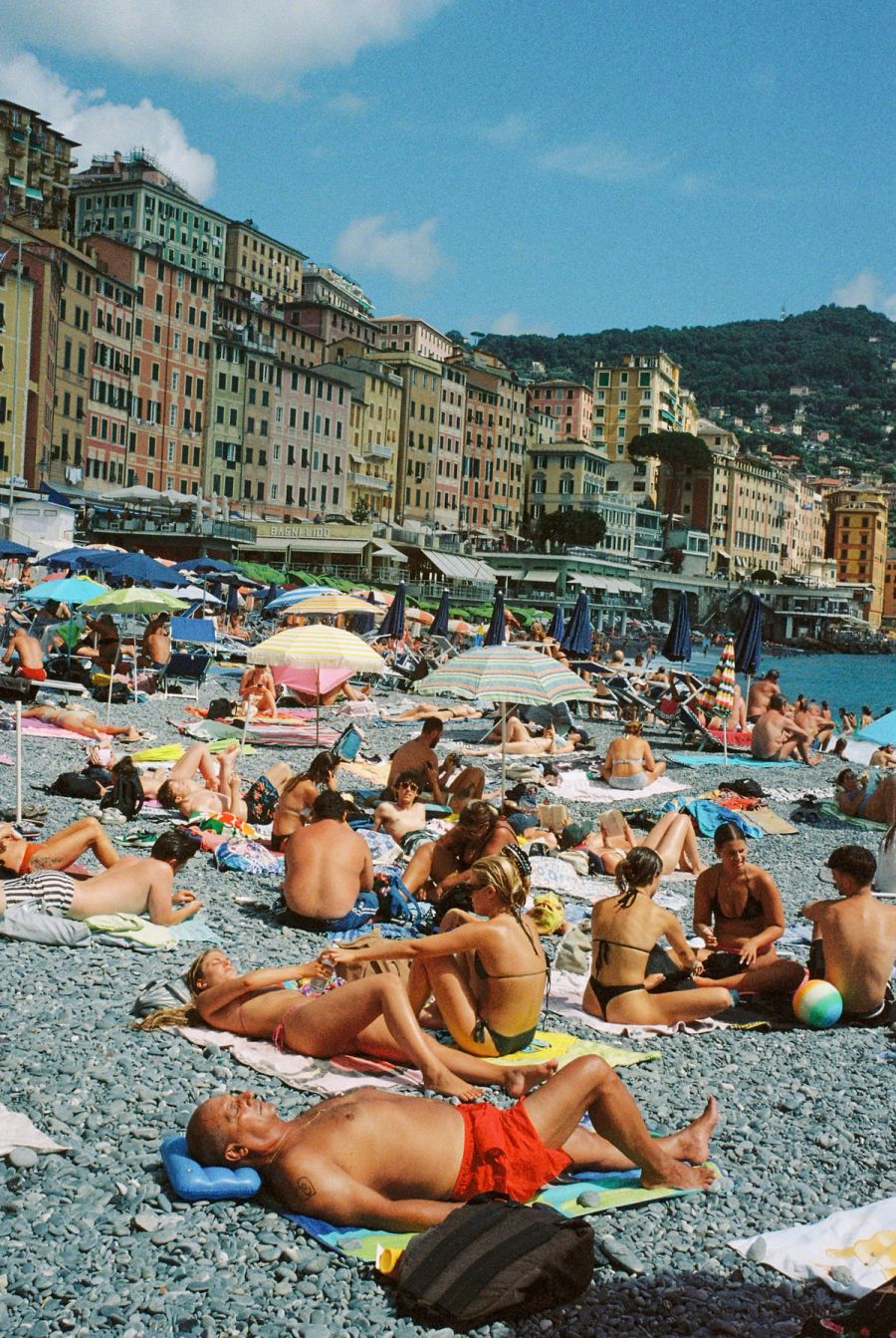 Sunbathers on a crowded stone beach beneath ochre facades on the Italian coast. Film photograph by Nicolò Pudel