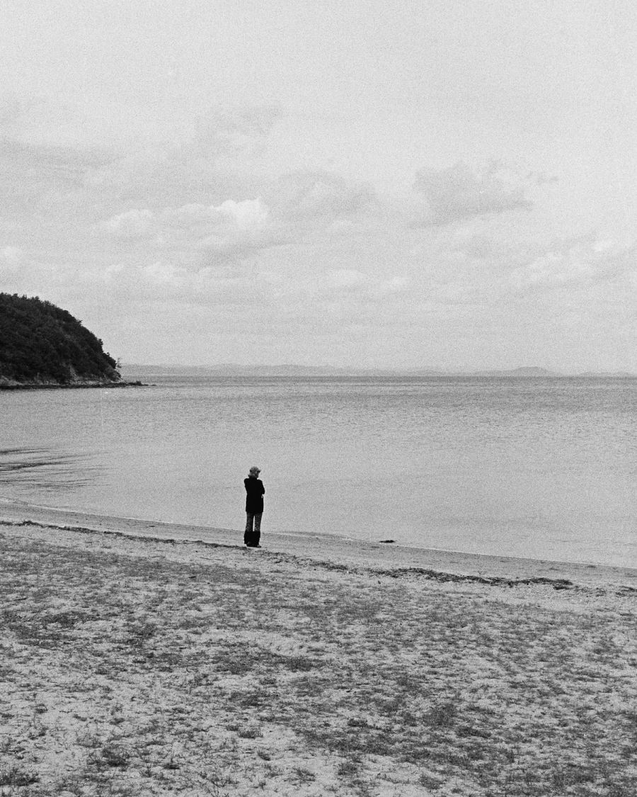 A person stands alone on a sandy beach facing calm water with a forested headland visible in the distance, film photograph by Nicolò Pudel.