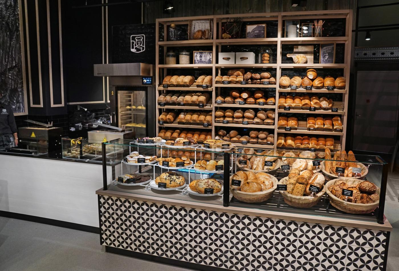 Ambachtelijke broodpresentatie bij Echte Bakker Hagedoren in Beek, met versgebakken broden en broodjes in een stijlvol interieur met hout, zwart-witte tegels en zicht op de oven.