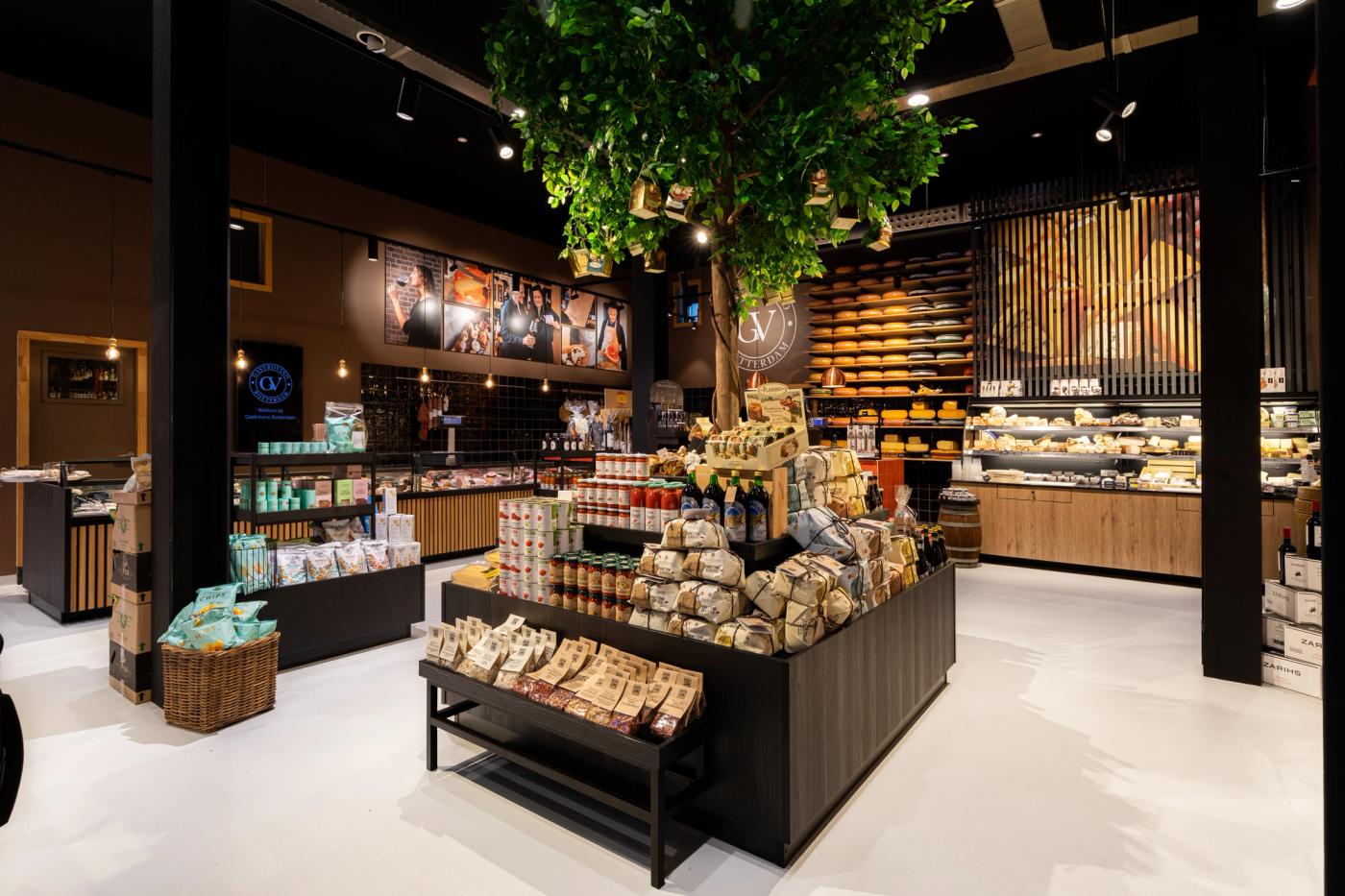 Luxe winkelinterieur van Gastrovino Rotterdam, ontworpen en gebouwd door Infrashop, met maatwerk kaaspresentatie, delicatessen, sfeervolle verlichting en opvallende groene boom als blikvanger.