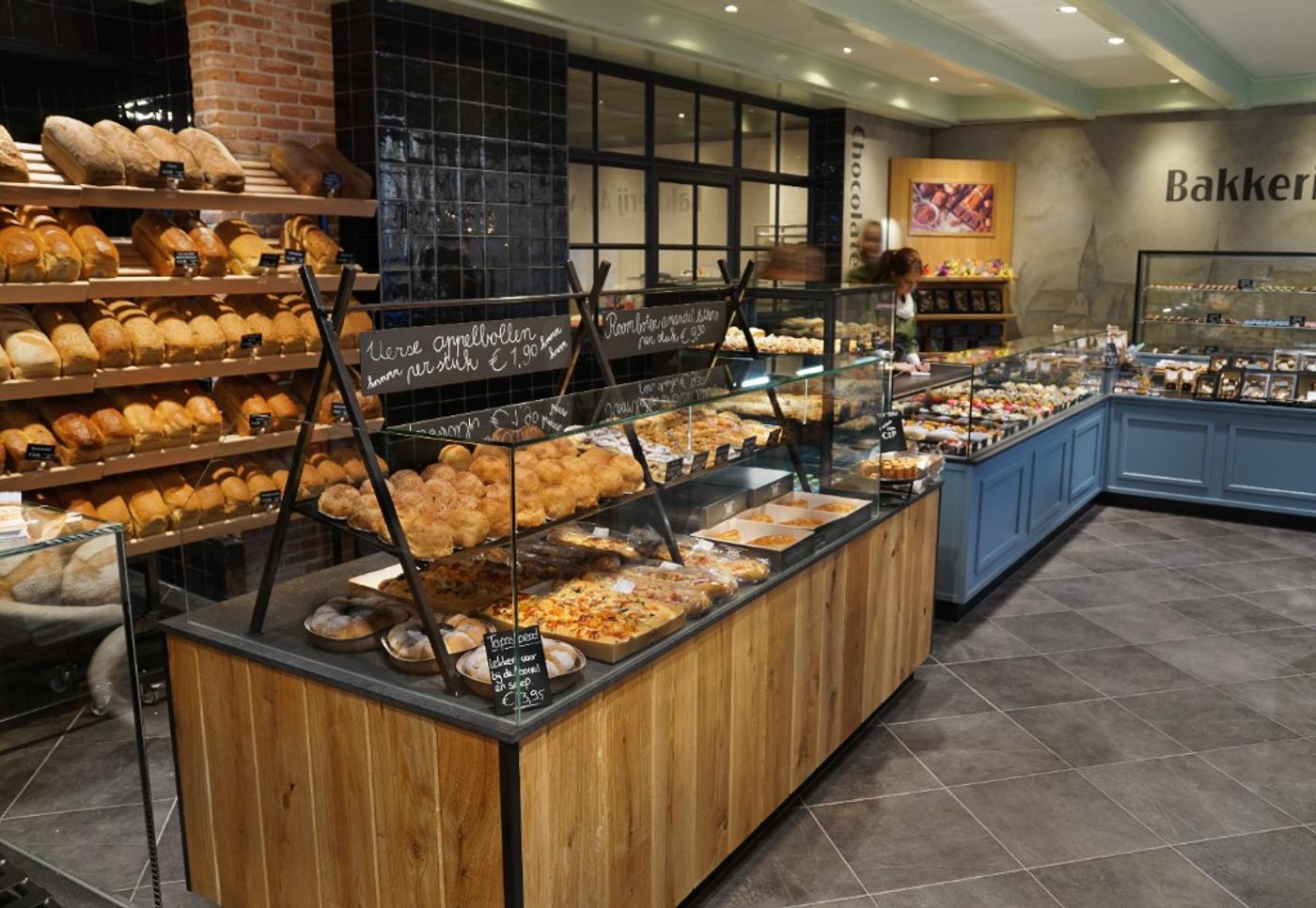 Interieur van een moderne ambachtelijke bakkerij met houten broodpresentatie, gebaksvitrines, blauwe toonbank en warme industriële details.